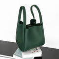 Medium Song Bag in emerald green pebbled leather with a structured bucket design and dual top handles, resting on books.