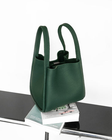 Medium Song Bag in emerald green pebbled leather with a structured bucket design and dual top handles, resting on books.