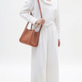 Model wearing a white shirt and pants, holding a medium-sized brown pebbled leather song bag with a shoulder strap.