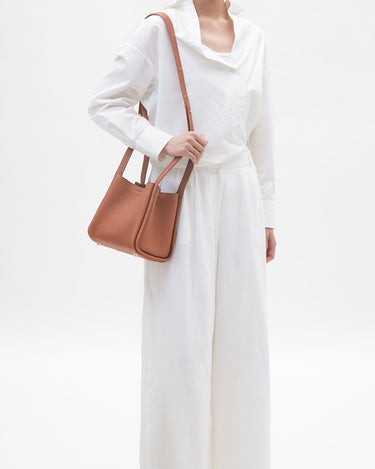 Model wearing a white shirt and pants, holding a medium-sized brown pebbled leather song bag with a shoulder strap.