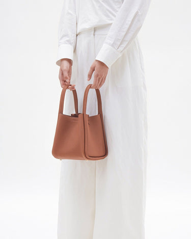 Medium Song Bag in rich, pebbled leather, held by hands in a white shirt and pants.