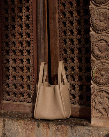 Medium Song Bag in taupe full-grain leather with dual handles against carved wood panels.