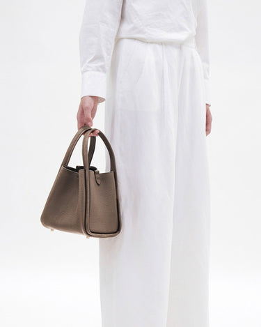 Medium Song Bag in taupe full-grain leather with textured finish and rounded handles, worn with white linen attire.