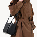 Medium Song Bag in Eco Onyx: black vegan leather bucket bag with gold hardware, worn by model in brown coat.