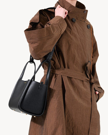 Medium Song Bag in Eco Onyx: black vegan leather bucket bag with gold hardware, worn by model in brown coat.