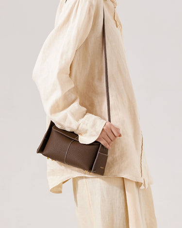Brown Mini Drippy Roof Bag in textured leather with white stitching, worn by a person in a cream linen outfit.