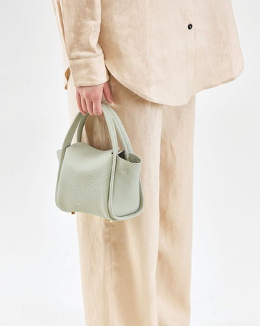 Mini Song Bag in Jade pebble leather, a compact bucket bag with gold hardware, held by a person in beige linen clothing.