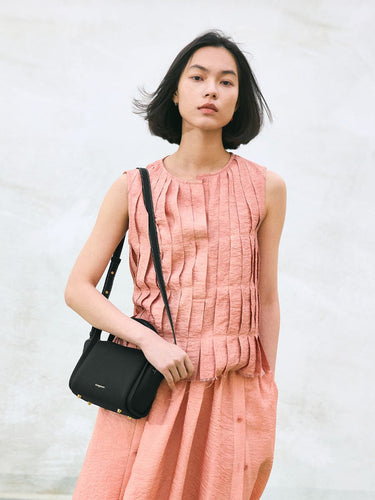 Mini Song Bag in Onyx black pebbled leather with gold hardware, worn by a woman in a peach pleated dress.