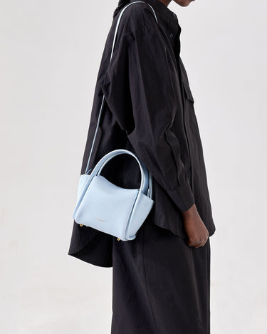 Sky blue pebbled leather Mini Song Bag with gold hardware, carried by a model in a black oversized shirt.