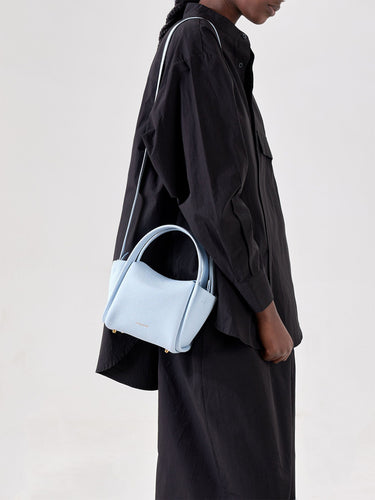 Sky blue pebbled leather Mini Song Bag with gold hardware, carried by a model in a black oversized shirt.