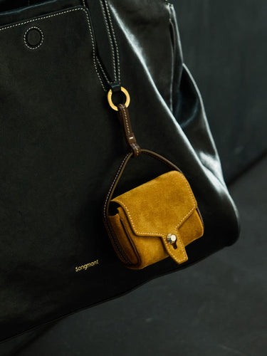 Ochre suede Mini Yore AirPods Case with snap closure, attached to a black leather Songmont handbag.