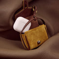 Ochre Mini Yore AirPods Case with white AirPods inside, made of suede and leather with metal clasp.