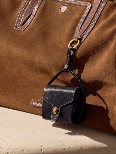 Mini Yore AirPods case in navy leather with gold clasp, attached to a tan suede tote bag.