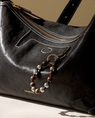 Black leather handbag featuring a sandalwood and silver bead charm attached to the side, with embossed "Songmont" logo.