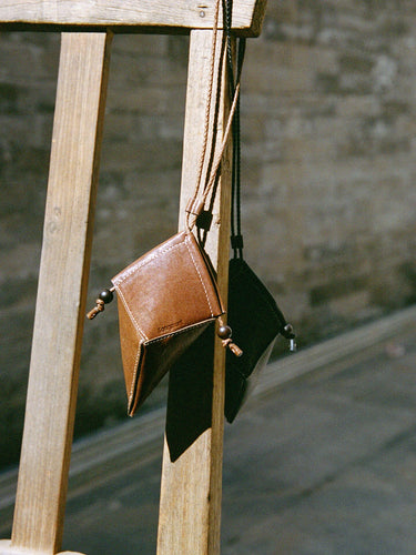 Prism Airpods Case in Sandal leather, featuring a geometric design and braided strap, hanging from a wooden structure.