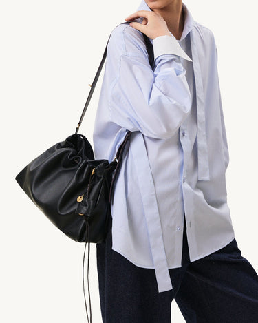 Black leather small gather bag set with gold charm, worn by a person in a pinstripe shirt.