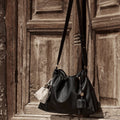 Small Gather Bag Set in Onyx. Black Nappa leather shoulder bag with a cream pouch, hanging on a rustic wooden door.