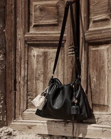 Small Gather Bag Set in Onyx. Black Nappa leather shoulder bag with a cream pouch, hanging on a rustic wooden door.