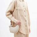 Ivory Small Luna Bag: pebbled leather, curved shape, adjustable strap. Model wears matching beige linen.