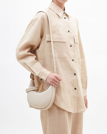 Ivory Small Luna Bag: pebbled leather, curved shape, adjustable strap. Model wears matching beige linen.