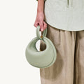 Sage green pebble-grain Small Luna Bag with twisted top handle and gold hardware, paired with a textured light green blouse and beige pants.
