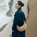 Medium Luna Hobo Bag in Dune Suede, crafted from cow suede leather and full-grain leather, worn by a woman on a beach.