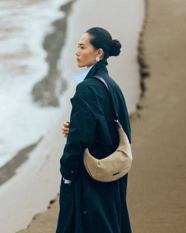 Medium Luna Hobo Bag in Dune Suede, crafted from cow suede leather and full-grain leather, worn by a woman on a beach.