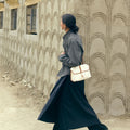 Small Shan Satchel Bag in cream canvas with tan leather trim, worn by a woman in a grey shirt and black pants walking by a textured wall.