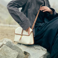 Small Shan Satchel Bag in canvas with tan leather trim, worn by a person sitting on rocks.