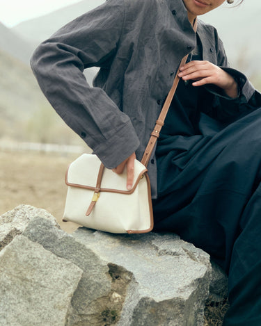 Small Shan Satchel Bag in canvas with tan leather trim, worn by a person sitting on rocks.