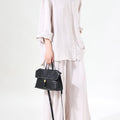Black Small Shan Satchel Bag with textured leather and gold hardware, worn by a model in linen clothing.