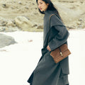 Model wearing a grey coat and carrying a small brown leather satchel bag.