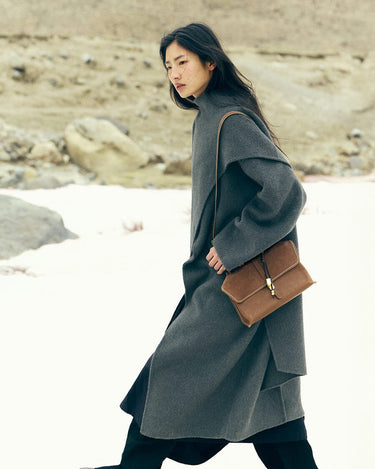 Model wearing a grey coat and carrying a small brown leather satchel bag.