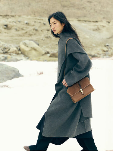 Model wearing a grey coat and carrying a small brown leather satchel bag.