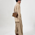 Model wearing tan shirt and pants, carrying a small brown leather satchel bag with a gold buckle.