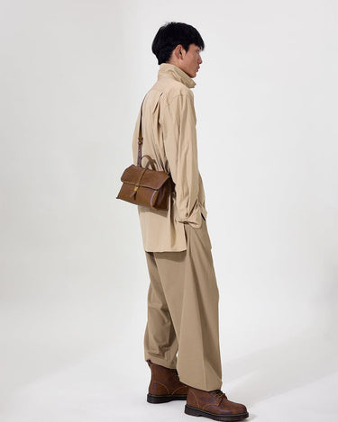 Model wearing tan shirt and pants, carrying a small brown leather satchel bag with a gold buckle.