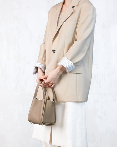A person holds a small taupe pebble-grain leather Songmont bag, wearing a cream blazer and white pants.