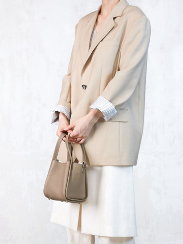 A person holds a small taupe pebble-grain leather Songmont bag, wearing a cream blazer and white pants.