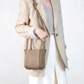 Woman wearing a beige blazer and white pants, holding a small taupe-colored pebbled leather Song bag.