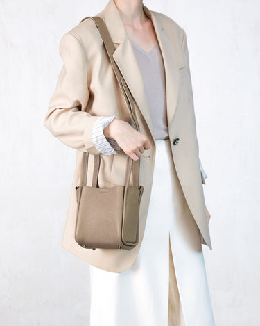 Woman wearing a beige blazer and white pants, holding a small taupe-colored pebbled leather Song bag.