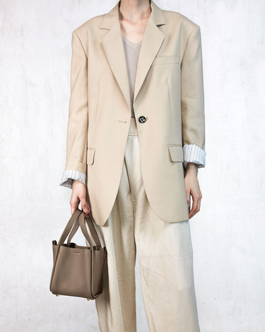 Small Song Bag in taupe pebbled leather, styled with a beige blazer and pants.