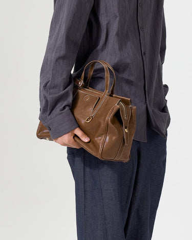 Brown Small Yore Duffle Bag in Sandal leather, white stitching, gold hardware. Carried by person in grey shirt.