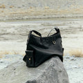Small Yore Hobo Bag in Onyx leather, featuring adjustable strap and exterior pocket, rests on a rock outdoors.